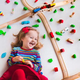 Child playing with wooden train, rails and cars. Toy railroad for kids. Educational toys for preschool and kindergarten children. Little girl at daycare. View from above, kid playing on the floor.